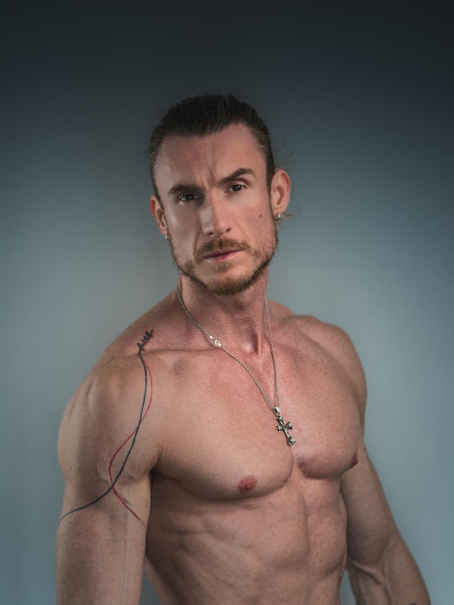 Adi Gheo in a shirtless athletic casting portrait against a blue-grey background