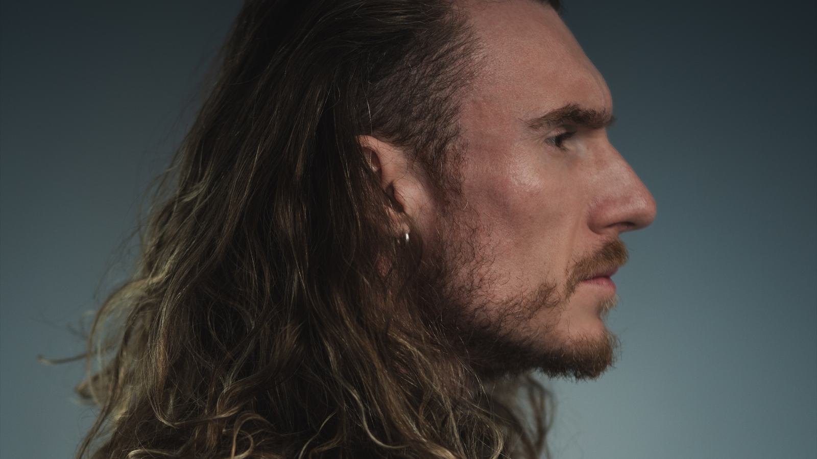 Side profile of Adi Gheo with long hair against a blue-grey background, showreel link image